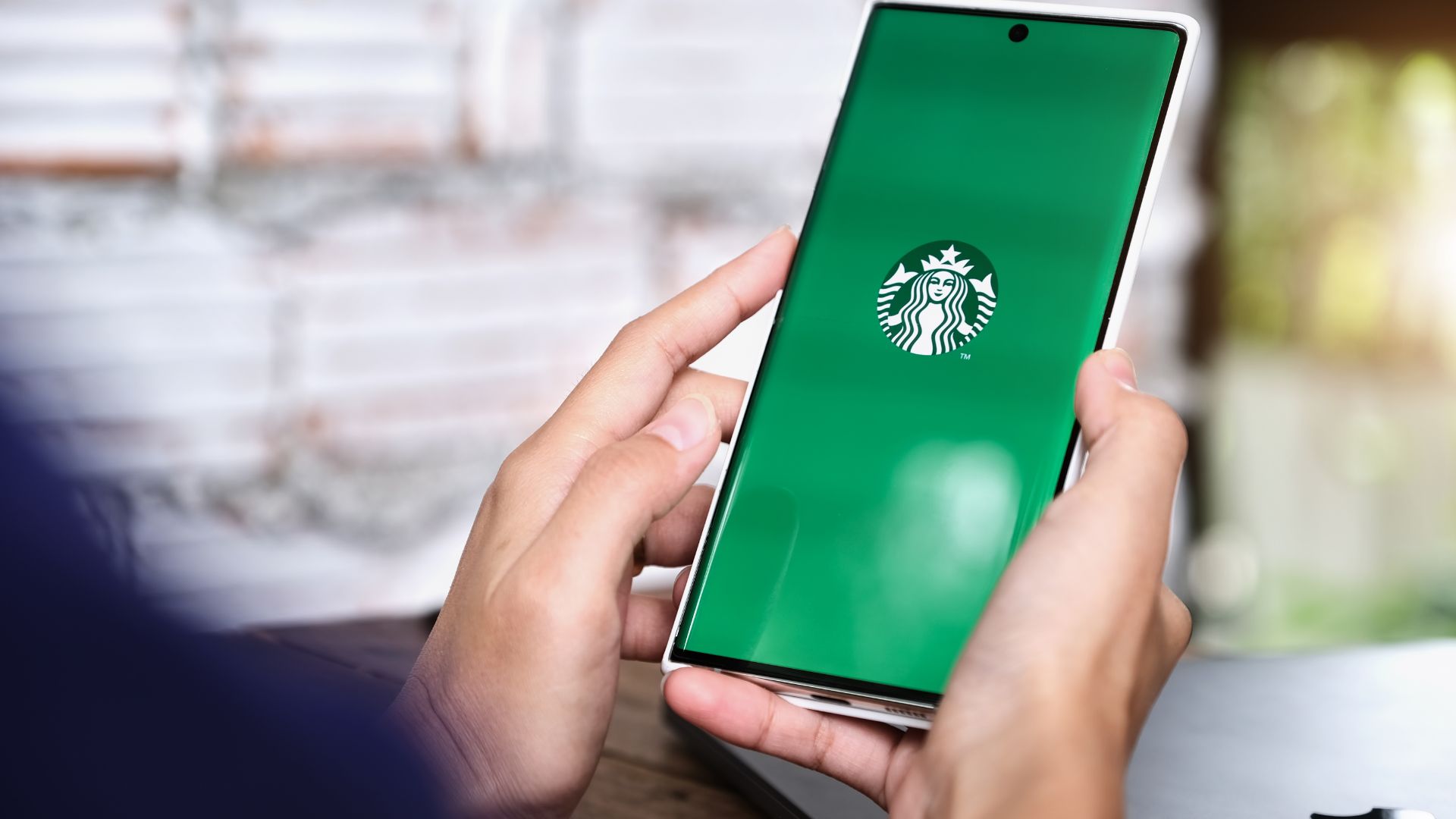 Hands holding a smartphone displaying the Starbucks app with a green screen and logo, showcasing mobile integration